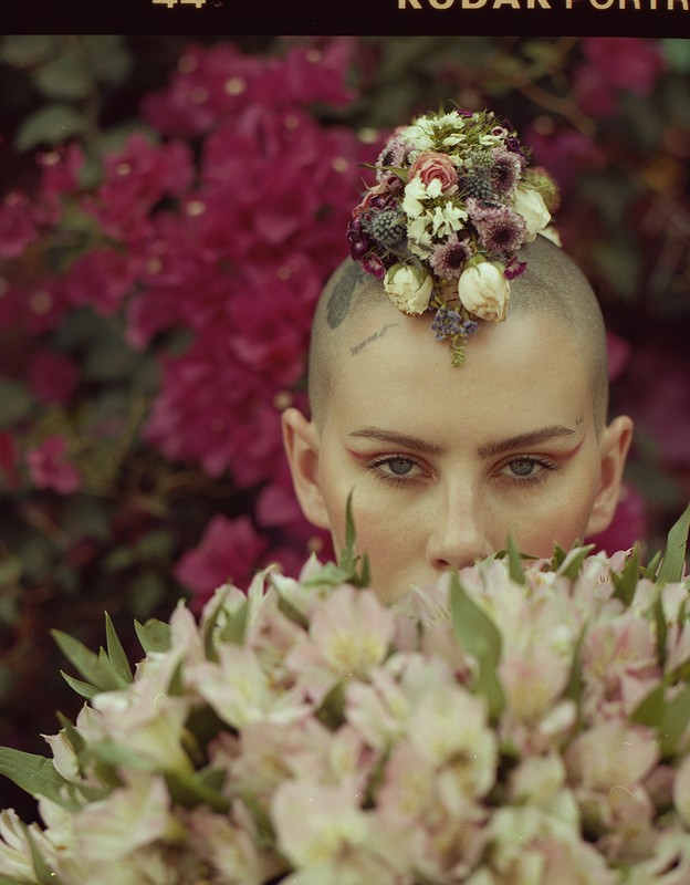Close-up portrait of a bald adult woman with floral headpiece, standing in front of magenta flowers and holding bouquet of light pink lilies.