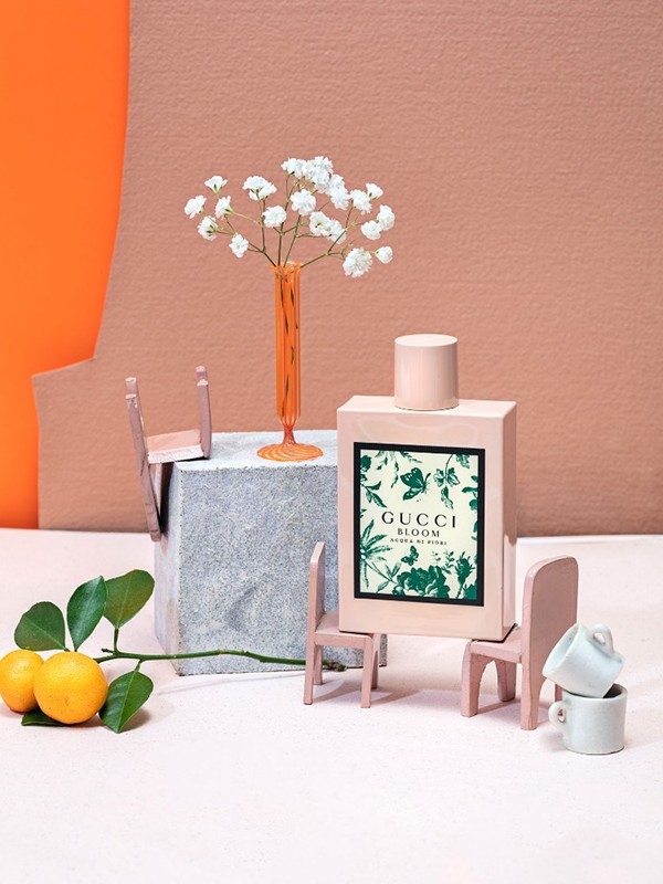 Gucci Bloom perfume bottle surrounded by miniature chairs, lemons on a branch, and white flowers, set against orange and beige backdrops.