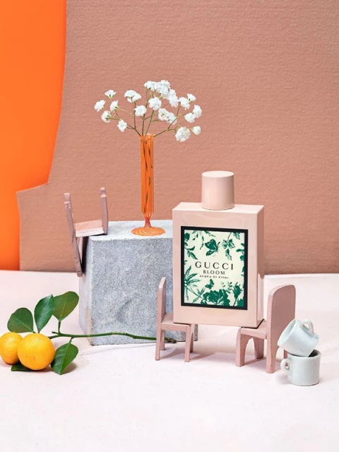 Gucci Bloom perfume bottle surrounded by miniature chairs, lemons on a branch, and white flowers, set against orange and beige backdrops.