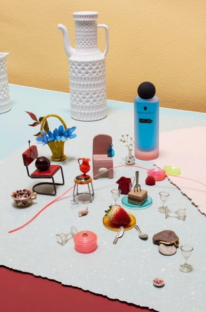 Still life of miniature objects, including furniture, dishware, a strawberry, flowers, and a perfume bottle, arranged on a textured surface.
