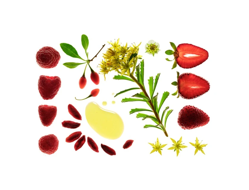 Overhead shot of raspberries, strawberries, yellow flowers, leaves, goji berries, and a pool of oil arranged on a white surface.