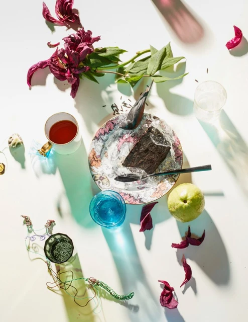Overhead shot of a dessert plate with cake, spoon, fruit, flower petals, tea cup, and decorative objects against a white backdrop.