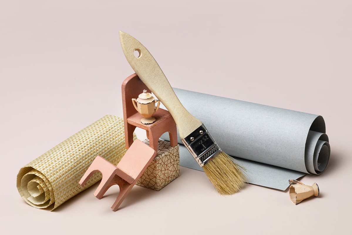 Still life shot: a paint brush, rolls of textured paper, and miniature wooden furniture on a light pink background.