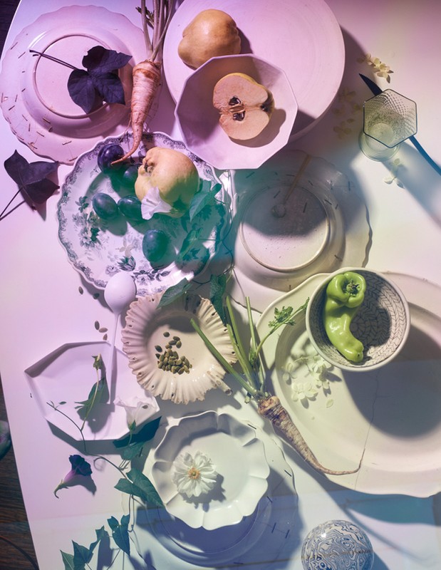 Overhead shot of white plates displaying pears, plums, parsley root, green peppers, capers, and flowers on a white table with purple lighting.