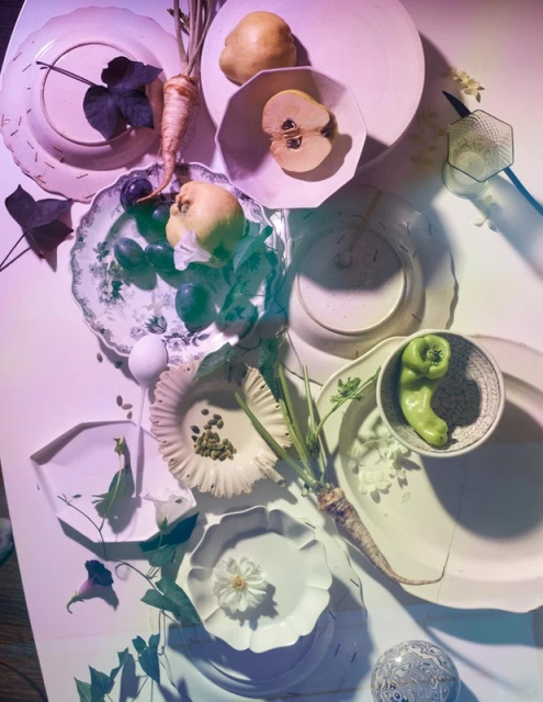 Overhead shot of white plates displaying pears, plums, parsley root, green peppers, capers, and flowers on a white table with purple lighting.