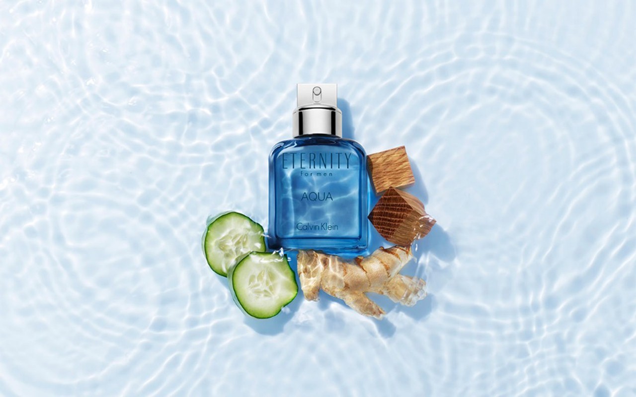 Eternity Aqua men's cologne by Calvin Klein on a rippled light-blue surface with cucumber slices and raw ginger accents, showcasing a fresh, aquatic fragrance.