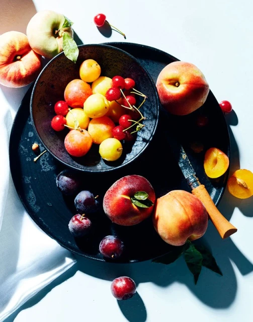 Overhead shot of a dark tray holding peaches, plums, apricots, and cherries next to a knife and white fabric with shadows.
