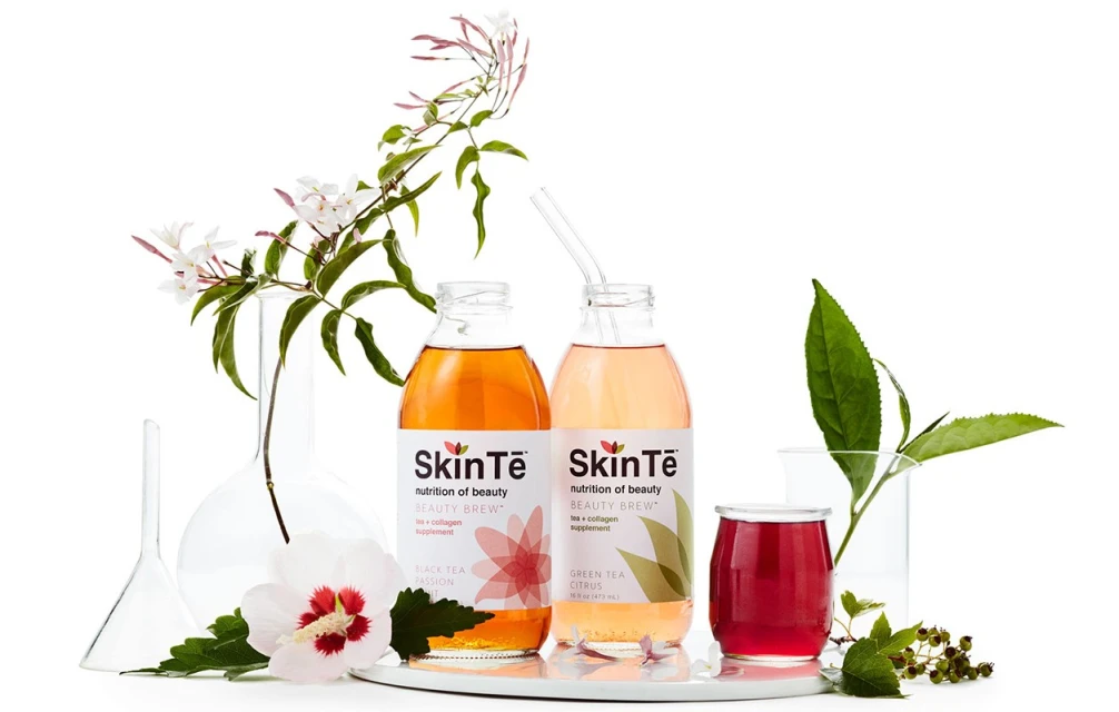 Two bottles of SkinTē Beauty Brew in Black Tea Passion and Green Tea Citrus flavors, with flowers and glassware against a white background.