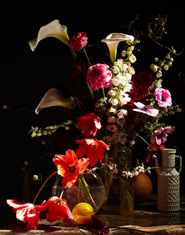 Dramatic still life of assorted flowers including tulips and calla lilies in glass vases on a dark marble surface against a black background.