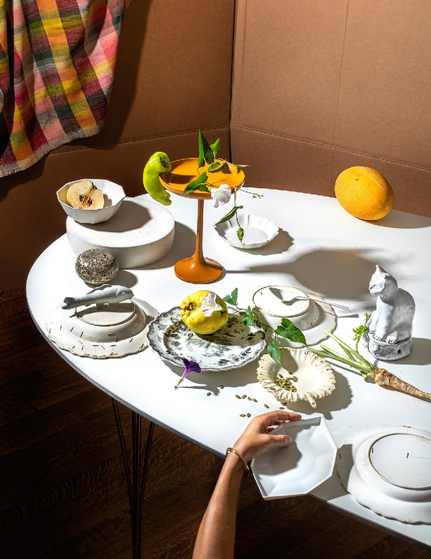 Still life featuring an arrangement of dishes, fruit, and decorative objects on a white table, with a backdrop of brown and patterned fabric.