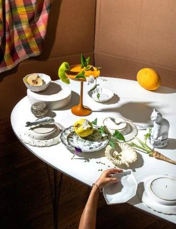 Still life featuring an arrangement of dishes, fruit, and decorative objects on a white table, with a backdrop of brown and patterned fabric.