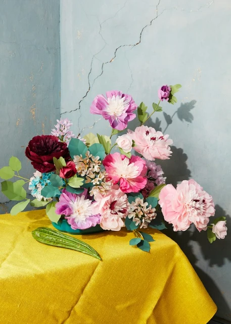 Still life photography of a vibrant floral arrangement with pink and red flowers in a turquoise vase on a yellow tablecloth.