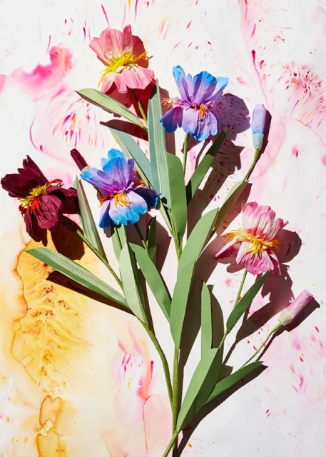 Artistic arrangement of colorful paper flowers with green stems on an abstract background with pink and yellow paint splashes.