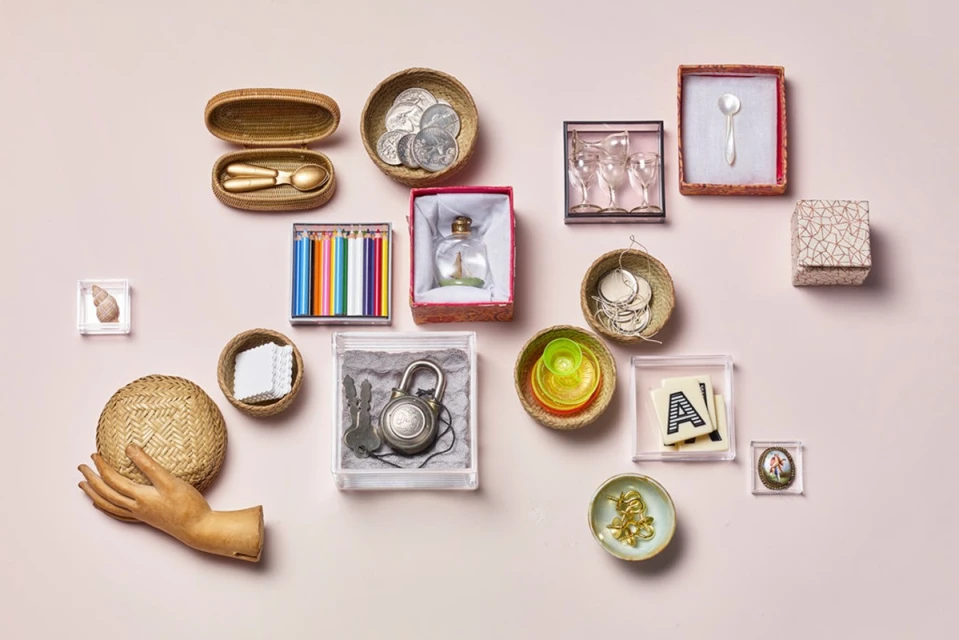 Arrangement of curated vintage objects in clear boxes and woven baskets, displayed on a pastel pink surface, studio lighting.