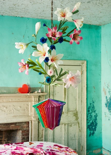 Hanging floral arrangement featuring lilies and paper art in a distressed, turquoise-painted room with aged furniture.