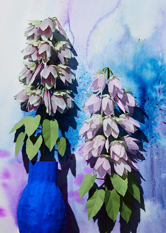 Still life of purple bellflowers with green leaves arranged in a blue vase against a textured, colorful watercolor background.
