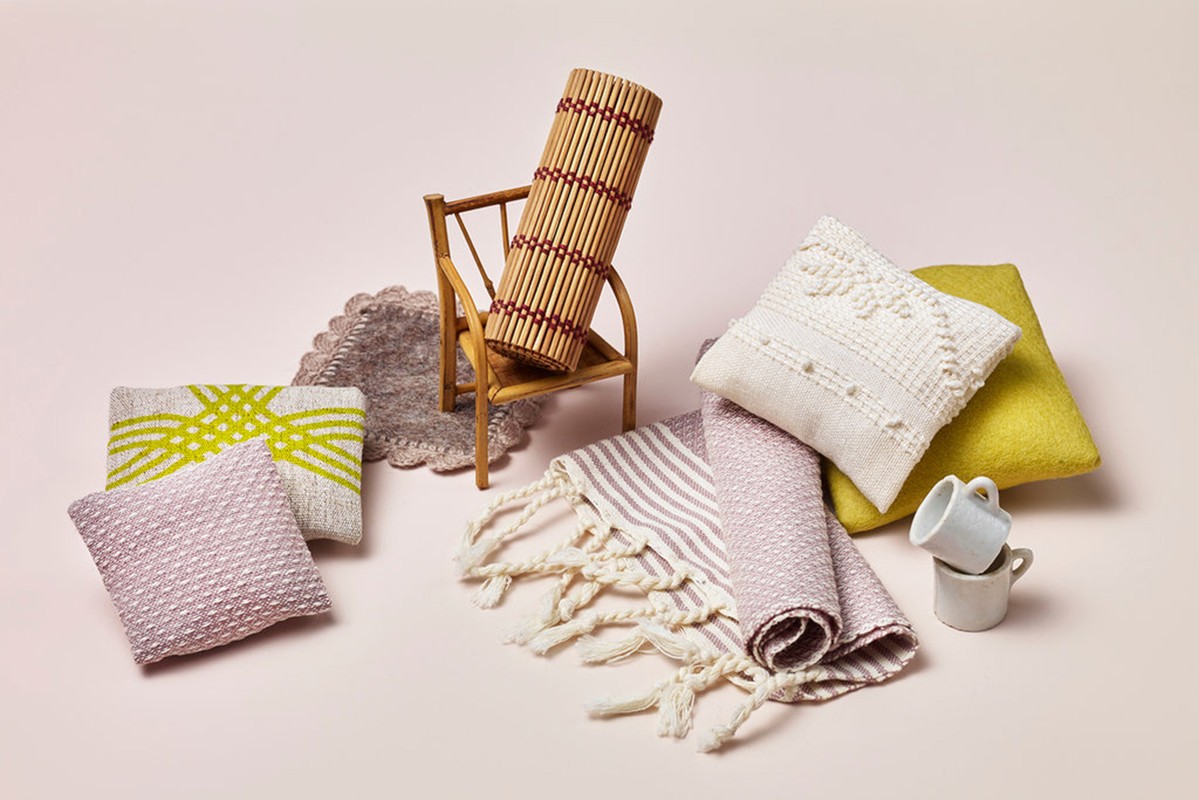 Still life of pillows, a rug, a small chair with a cushion, mugs, and a towel on a light pink background, creating a cozy scene.