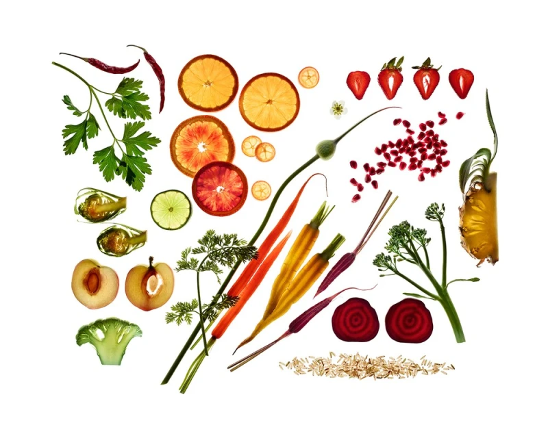 Overhead shot of sliced fruits and vegetables, including oranges, strawberries, plums, carrots, broccoli, beets, and pineapple on a white background.