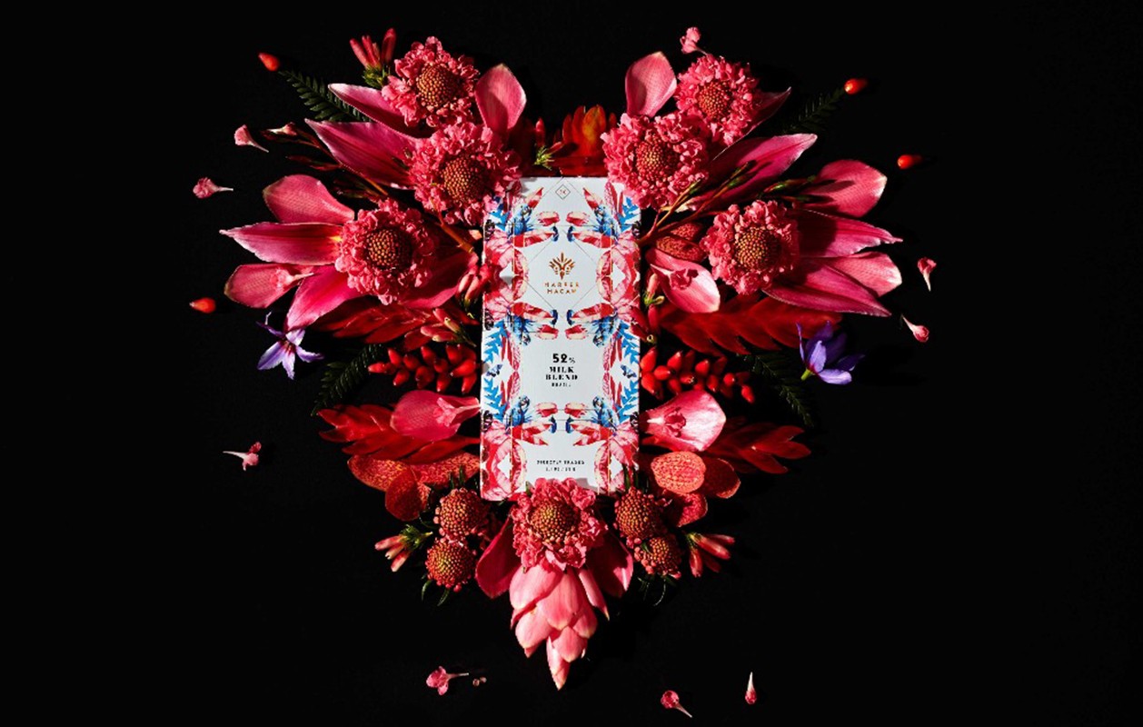 Overhead shot of a Tavalon Cocoa chocolate bar surrounded by bright pink and red flowers against a black background.