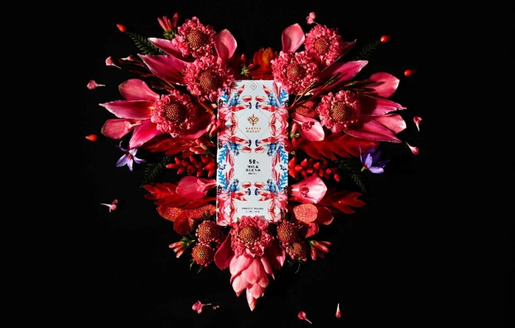 Overhead shot of a Tavalon Cocoa chocolate bar surrounded by bright pink and red flowers against a black background.