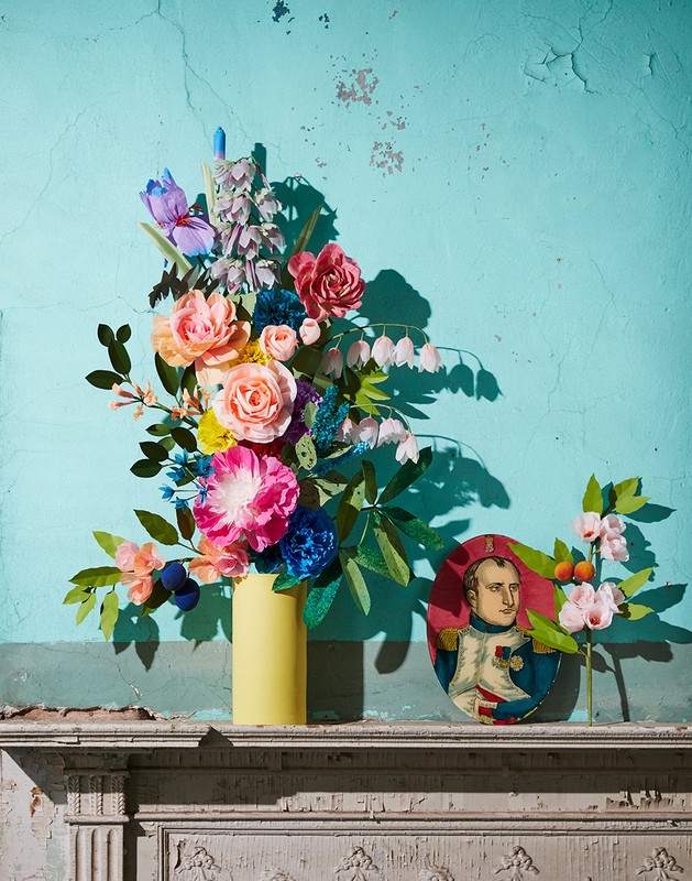 Paper flower arrangement in a yellow vase sits on a mantelpiece with a portrait against a turquoise wall, captured in a studio setting.
