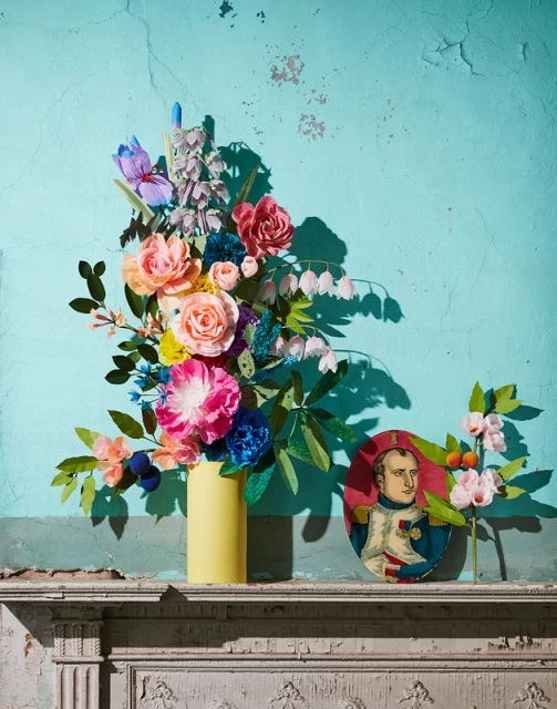 Paper flower arrangement in a yellow vase sits on a mantelpiece with a portrait against a turquoise wall, captured in a studio setting.