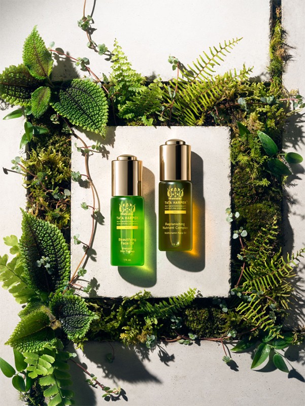 Two Tata Harper skincare bottles with gold caps, arranged on a square of concrete, surrounded by moss and lush green ferns and leaves.
