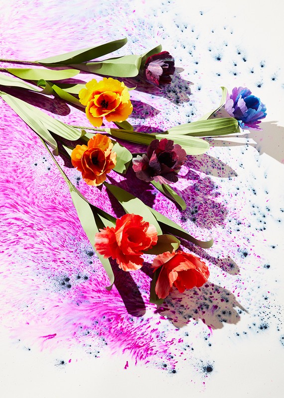 Floral flat lay with vibrant paper flowers in red, yellow, purple, and blue, set against a splatter-painted background of pink and white.