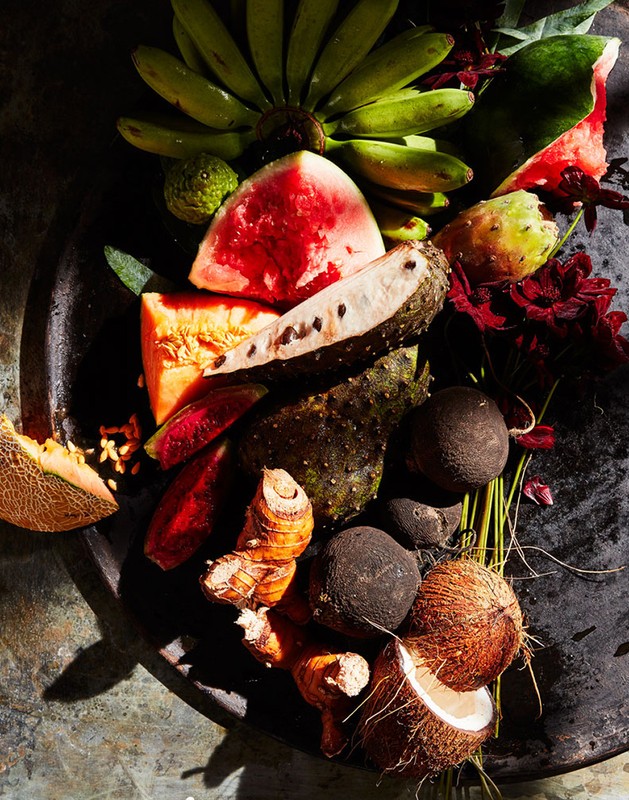 Overhead shot of fresh tropical fruits including bananas, melons, and coconuts arranged on a dark textured surface in bright lighting.