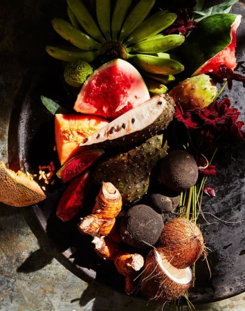 Overhead shot of fresh tropical fruits including bananas, melons, and coconuts arranged on a dark textured surface in bright lighting.