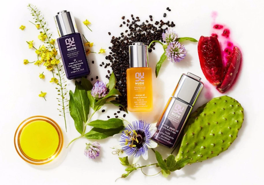 Three Nude Progenius skincare bottles of various colors are surrounded by flowers, seeds, and plants on a white surface.