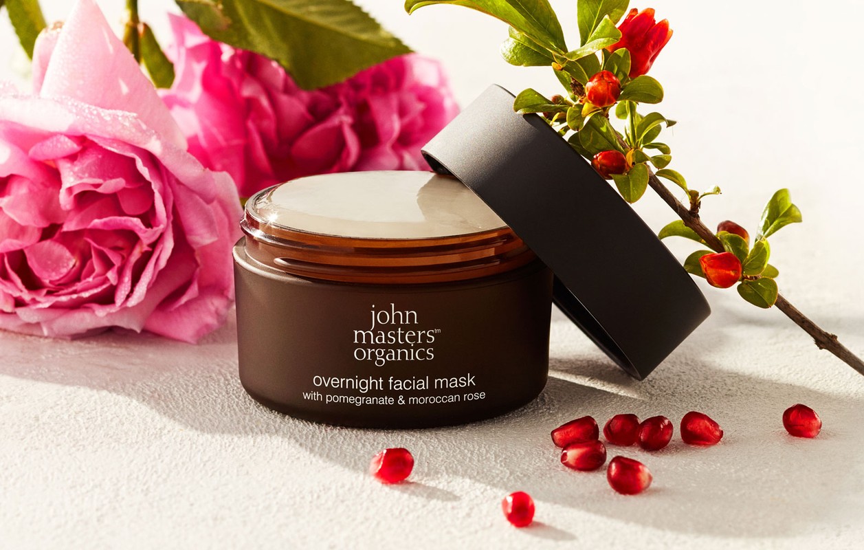 Brown jar of overnight facial mask with pomegranate and moroccan rose, accompanied by pink roses and branch of pomegranate.