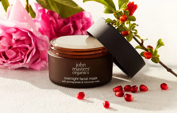 Brown jar of overnight facial mask with pomegranate and moroccan rose, accompanied by pink roses and branch of pomegranate.