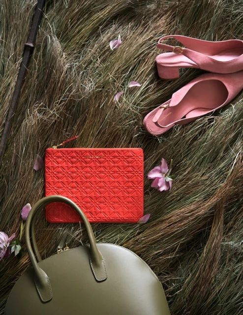 Fashion still life featuring an olive green satchel, red clutch, and pink slingback shoes arranged on a bed of straw with floral accents.