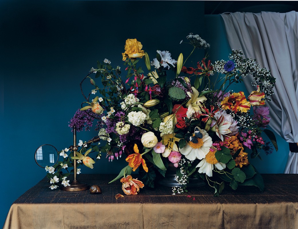 Still life of assorted flowers including roses and tulips in a vase on a table with a magnifying glass and snail shell.