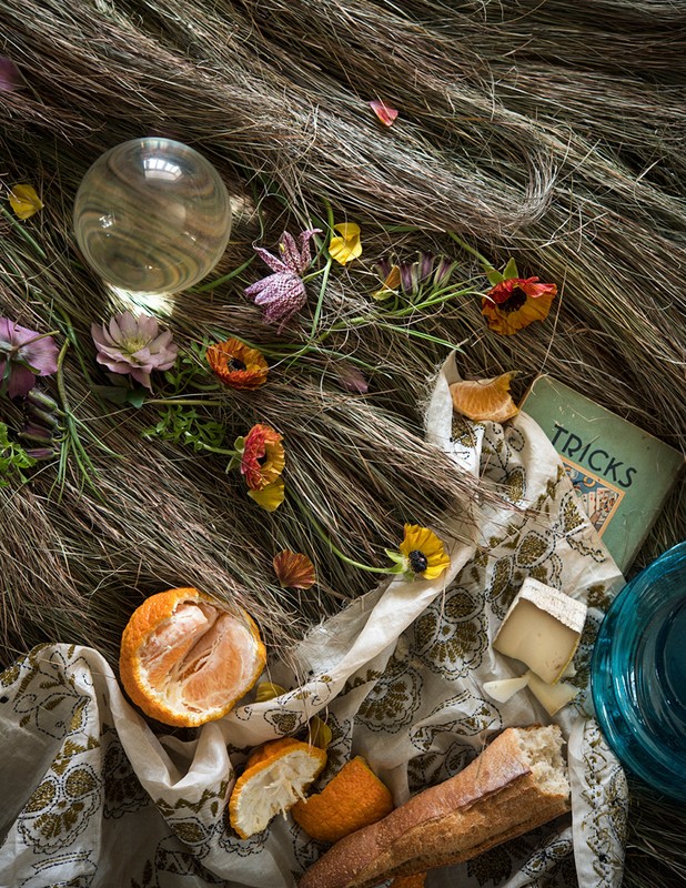 Still life photo featuring oranges, baguette, brie, flowers, and a glass orb arranged on a patterned cloth over brown grass. 