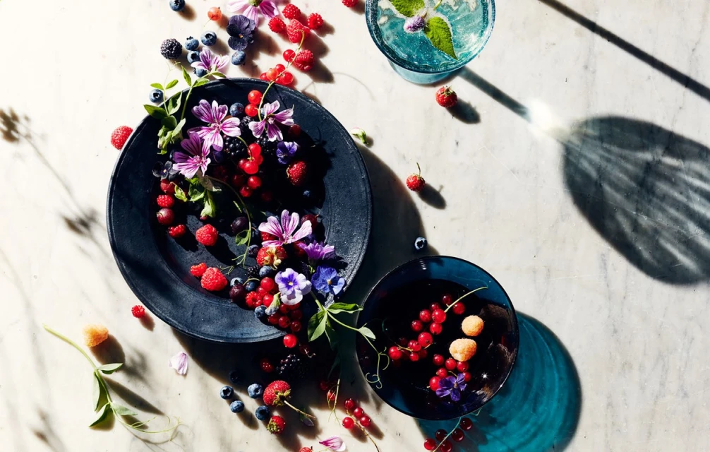 Overhead shot of assorted berries and flowers in black and blue bowls on a marble surface, with shadows and a refreshing beverage.
