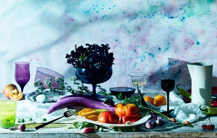 Still life photography of fresh vegetables and fruits arranged on a rustic wooden table against a mottled blue and white background.