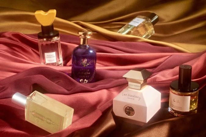 Several luxury perfume bottles arranged on draped satin fabric in rich burgundy and gold tones styled by Livia Cetti.