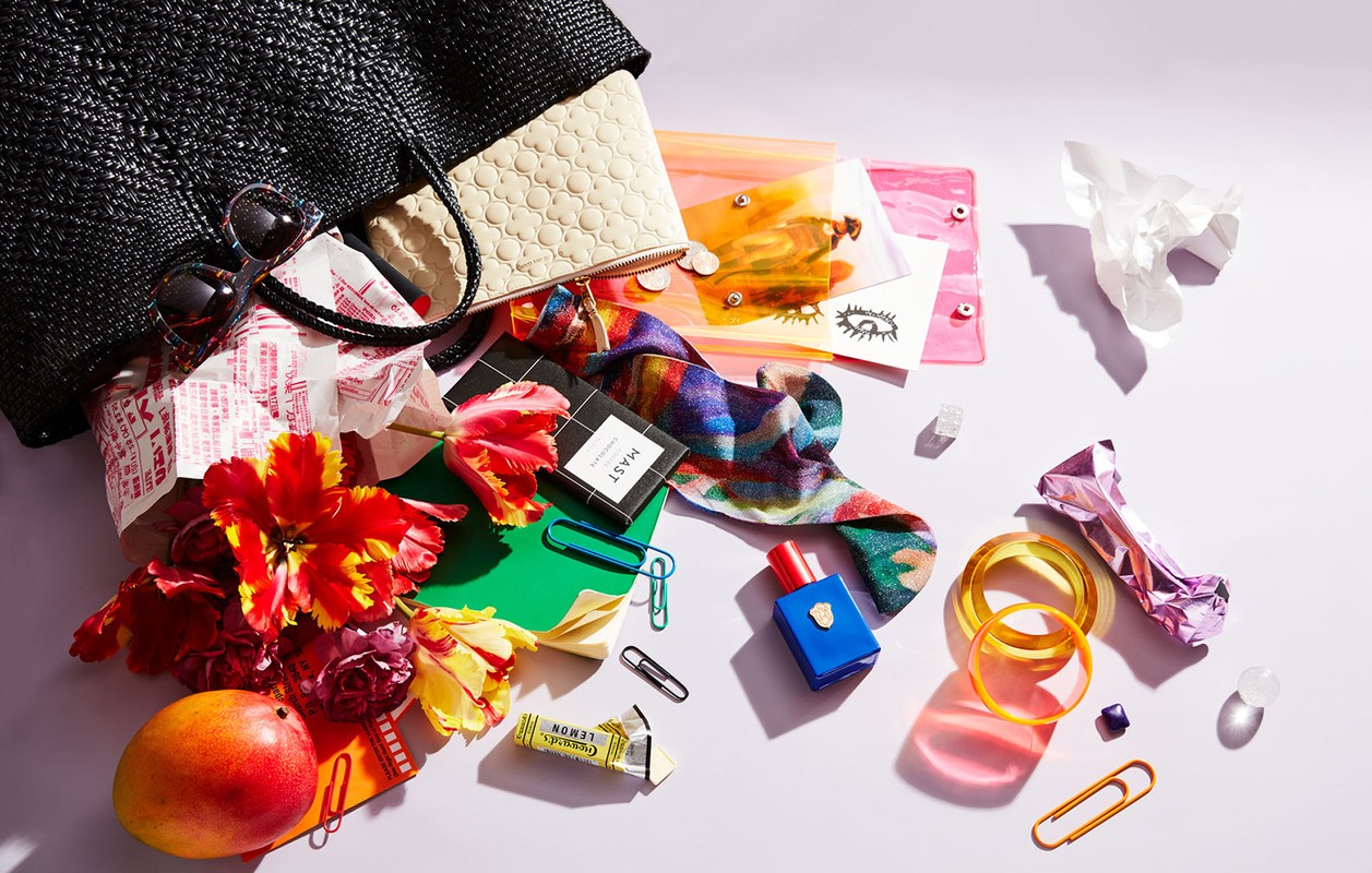 Still life of a black straw bag spilling out various colorful items, including flowers, a wallet, cosmetics, and accessories on a white surface.