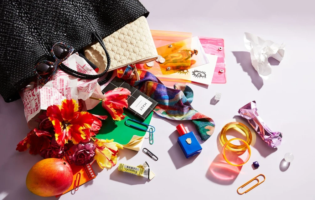 Still life of a black straw bag spilling out various colorful items, including flowers, a wallet, cosmetics, and accessories on a white surface.