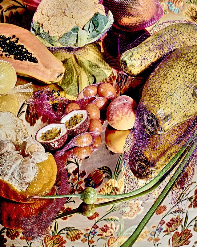 Still life shot of assorted fruits including papaya, passion fruit, melons, and cauliflower in netting on a floral-patterned surface.