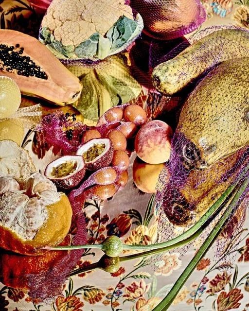 Still life shot of assorted fruits including papaya, passion fruit, melons, and cauliflower in netting on a floral-patterned surface.