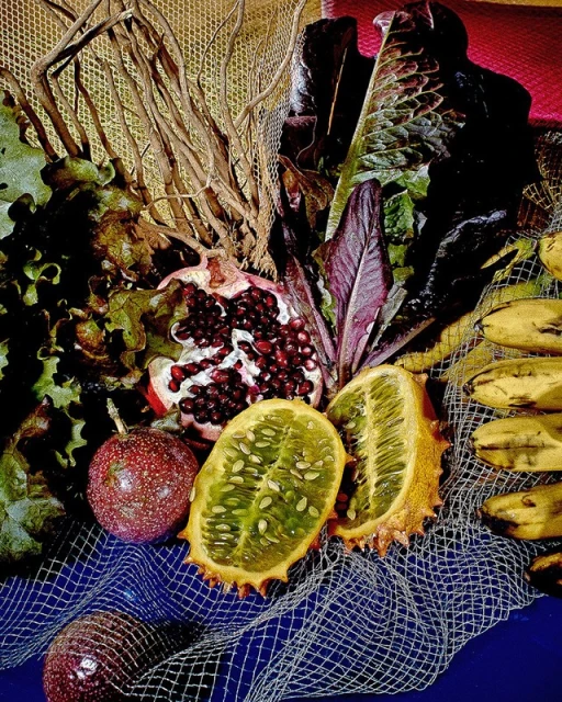 Still life shot with tropical fruits including horned melon, bananas, and passion fruit, plus pomegranate, lettuce, and branches on a blue background.