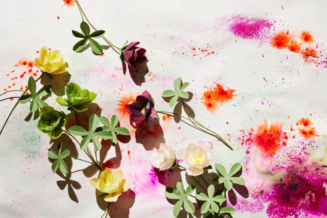 Artistic flat lay with paper flowers in yellow, green, burgundy and white, complemented by paint splatters in red, pink and turquoise.