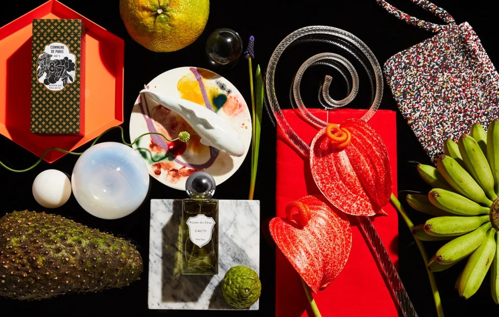 Still life featuring perfume, flowers, and various fruits arranged on a black surface with a mix of textures and vibrant colors.