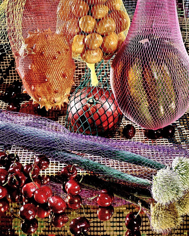 Still life composition with kiwano melons, plums, cherries, and apples in colorful mesh bags creating a vibrant, textured visual.