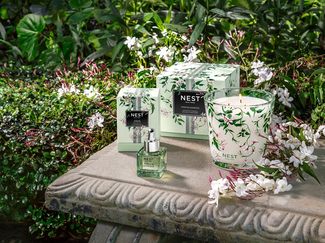 Nest New York Indian Jasmine scented candle and diffuser set on a stone surface outdoors amid lush green foliage and white jasmine flowers.