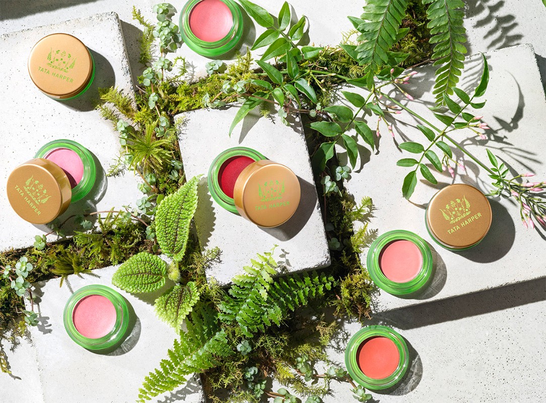 Overhead studio shot of Tata Harper blushes in green and gold containers arranged among lush green ferns and moss on concrete.