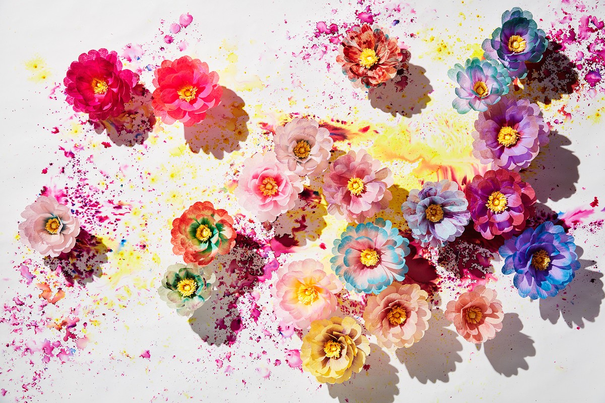 Overhead shot of colorful paper flowers with a yellow and magenta paint splatter on a white surface creating a vibrant floral arrangement.